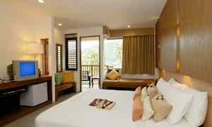 Hotellihuone, jossa on iso sänky, useita tyynyjä, televisio, istuinryhmä ja parveke, jolta näkyy palmuja ja vuoria. (Tekoälyn luoma teksti)