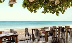 Ulkoterassi, jossa pöytiä ja tuoleja, sijaitsee rannalla ja taustalla näkyy meri. (Tekoälyn luoma teksti)