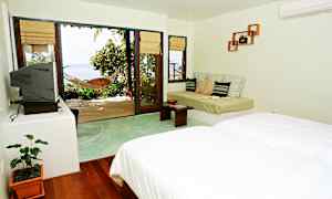 Valoisa hotellihuone, jossa on sänky, sohva ja suuri lasiovi, joka johtaa terassille riippukeinut ja näkymät merelle. (Tekoälyn luoma teksti)