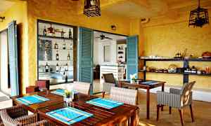 Huone, jossa on keltaiset seinät ja turkoosit ovet, sisustettu puupöydillä ja rottinkituoleilla. Hyllyillä näkyy pulloja ja tarjoiluastioita. (Tekoälyn luoma teksti)