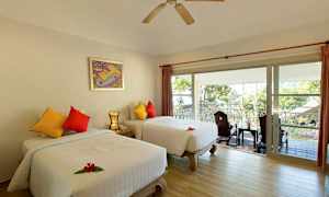 Hotellihuone, jossa on kaksi erillistä sänkyä, värikkäitä tyynyjä ja parveke, jolta avautuu näkymä puihin ja katolle. (Tekoälyn luoma teksti)