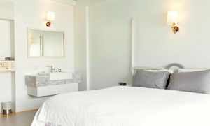 Vaalea hotellihuone, jossa on sänky harmailla tyynyillä ja pesuallas peilillä. (Tekoälyn luoma teksti)