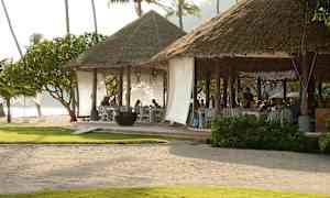 Rantaravintola, jossa on olkikatto ja avoimia istumapaikkoja, ympäröitynä vehreydellä ja palmuilla. (Tekoälyn luoma teksti)