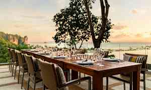 Kattaus pöydällä ja tuoleilla ulkona auringonlaskussa, taustalla meri ja puita. (Tekoälyn luoma teksti)