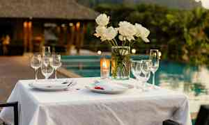 Pöytä, joka on katettu valkoisella liinalla, lautasilla, laseilla ja valkoisilla ruusuilla maljakossa, taustallaan uima-allasalue ja vehreä ympäristö. (Tekoälyn luoma teksti)