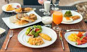 Aamiainen, jossa on erilaisia ruokalajeja, kuten munakas, salaatti, leivonnaiset, mehua ja hedelmiä, aseteltuna lautasille pöydälle. (Tekoälyn luoma teksti)