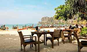 Ulkotarjoilualue, jossa pöytiä ja tuoleja rannalla, taustalla pitkähäntäveneitä ja kallioita rannikolla. (Tekoälyn luoma teksti)