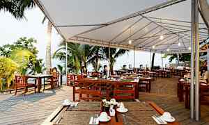 Ulkoterassi, jossa on puupöytiä ja -tuoleja valkoisen katoksen alla, ympäröity trooppisilla palmuilla. (Tekoälyn luoma teksti)