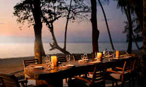 Pitkä pöytä ulkona terassilla rannalla, ympärillä puita ja näkymä merelle auringonlaskussa. Pöydälle on katettu lautasia, laseja ja kynttilälyhtyjä. (Tekoälyn luoma teksti)