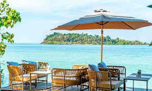 Ulkohuonekalut, joissa on tyynyjä, päivänvarjon alla, taustalla meri ja vihreä saari. (Tekoälyn luoma teksti)