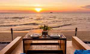 Kahdelle katettu pöytä, jossa on lautaset ja aterimet, sijaitsee meren rannalla auringonlaskun aikaan. (Tekoälyn luoma teksti)
