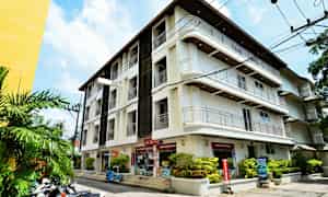 Kuva valkoisesta rakennuksesta, jossa on useita kerroksia. Rakennuksen ympärillä on vihreitä kasveja ja vieressä on moottoripyöriä pysäköitynä. (Tekoälyn luoma teksti)