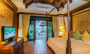 Makuuhuone, jossa on puupaneeleja, ja näkymä terassille sekä merelle. Huoneessa on sänky värikkäillä tyynyillä, TV-kaappi ja lamppuja. (Tekoälyn luoma teksti)