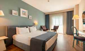 Hotellihuone, jossa on parivuode, kaksi nojatuolia pöydän ääressä, kirjoituspöytä ja televisio. (Tekoälyn luoma teksti)