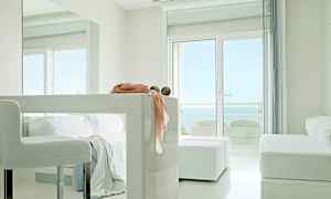 Valoisa, moderni huone, jossa on valkoisia huonekaluja, suuret ikkunat merinäköalalla ja koristeellisia yksityiskohtia pöydällä. (Tekoälyn luoma teksti)