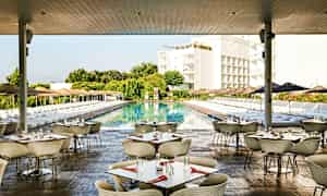 Kuva ulkoilmaravintolasta, jossa on pöytiä ja tuoleja, suuren uima-altaan vieressä. Altaan ympärillä on aurinkotuoleja ja aurinkovarjoja, taustalla vehreyttä ja rakennuksia. (Tekoälyn luoma teksti)