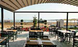 Ulkoterassilla pöytiä ja tuoleja, taustalla hiekkaranta ja meri. (Tekoälyn luoma teksti)