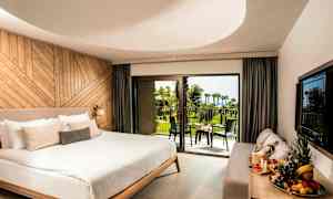 Moderni hotellihuone, jossa on suuri parivuode, sohva ja pöytä hedelmätarjottimen kera. Huoneessa on lasiliukuovet, jotka johtavat parvekkeelle, jolta on näkymä vehreään puutarhaan ja taustalla olevalle merelle. (Tekoälyn luoma teksti)