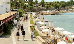 Kävelytie meren äärellä, jossa ihmisiä kävelee ja henkilöitä rentoutumassa aurinkotuoleilla varjojen alla. (Tekoälyn luoma teksti)