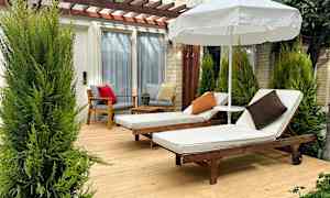 Kaksi aurinkotuolia, joissa on valkoisia patjoja ja tyynyjä, seisovat puisella terassilla varjon alla, ympärillä vihreitä kasveja. (Tekoälyn luoma teksti)