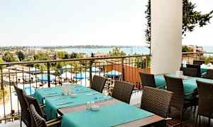 Näkymä ravintolasta, jossa katettuja pöytiä ja tuoleja, ulkoalue aurinkovarjoineen ja meri taustalla. (Tekoälyn luoma teksti)