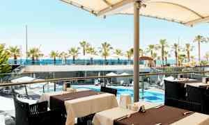 Näkymä ulkoilmaravintolasta kohti uima-allasaluetta, jossa on aurinkovarjoja ja palmuja sekä taustalla meri. (Tekoälyn luoma teksti)