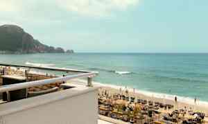 Kuva rannasta, jossa on aurinkotuoleja, aurinkovarjoja ja ihmisiä uimassa meressä. Taustalla näkyy kallioluoto. (Tekoälyn luoma teksti)