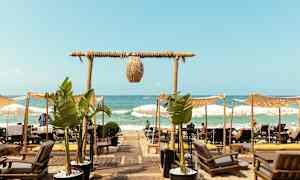 Näköala rannalle, jossa on aurinkotuoleja ja varjoja, ja polku palmuistutusten ohi johtaa merelle. (Tekoälyn luoma teksti)