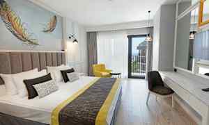 Hotellihuone, jossa on parisänky, keltainen nojatuoli, kirjoituspöytä ja peili sekä parveke merinäköalalla. (Tekoälyn luoma teksti)