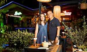 Kaksi henkilöä katetun pöydän ääressä, jossa on kakku ja kynttilöitä, ravintolassa vihreän kasvuston ja valaisimien joukossa. (Tekoälyn luoma teksti)