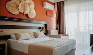 Hotellihuone, jossa oranssi seinä, parivuode kahdella tyynyllä sekä koriste-esineitä sängyn yläpuolella. (Tekoälyn luoma teksti)