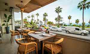 Kuvassa on ravintola, jolla on näkymä kadulle, jolla on pysäköityjä ja ohikulkevia autoja sekä palmuja taustalla. (Tekoälyn luoma teksti)