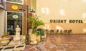 Rakennus, jonka vaalealla seinällä on "Orient Hotel" -kyltti, pienellä patiolla kalusteina tuoleja, pöytiä, kukkaruukkuja ja patsas. (Tekoälyn luoma teksti)