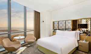 Kuva hotellihuoneesta, jossa on iso sänky, kaksi tuolia, pöytä ja ikkuna näkymällä. (Tekoälyn luoma teksti)