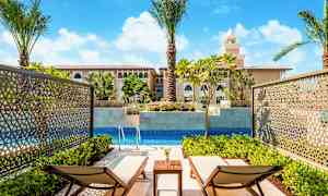 Kaksi aurinkotuolia yksityisen altaan äärellä, ympärillä vihreitä kasveja ja näkymä rakennukseen, jossa on välimerellisiä arkkitehtonisia elementtejä. (Tekoälyn luoma teksti)