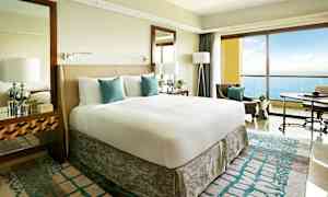 Moderni hotellihuone, jossa on suuri sänky, valkoiset ja turkoosit vuodevaatteet sekä parveke merinäköalalla. (Tekoälyn luoma teksti)