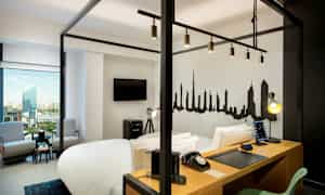 Moderni hotellihuone, jossa on neljän pylvässänky, työpöytä, televisio, istumaryhmä ja seinälle maalattu kaupunkimaisema. (Tekoälyn luoma teksti)