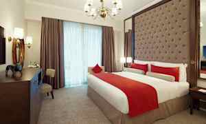 Tyylikkäästi sisustettu hotellihuone, jossa on suuri sänky, valkoiset ja punaiset vuodevaatteet. Huoneessa on pehmustettu sängynpääty, kattokruunu ja suuret ikkunat verhoilla. (Tekoälyn luoma teksti)