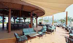 Ulkobaari, jossa istuimia ja puukatettu alue, ympärillä palmuja ja kaupungin siluetti taustalla. (Tekoälyn luoma teksti)