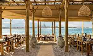 Ravintola rannalla, jossa puiset pöydät ja tuolit, merinäkymä ja kaupungin siluetti taustalla. (Tekoälyn luoma teksti)