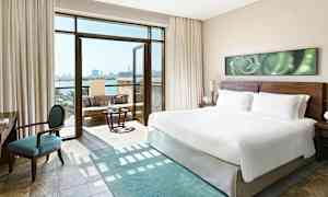 Moderni hotellihuone, jossa on suuri king-size-vuode, istuinryhmä ja avoin parveke merinäköalalla. (Tekoälyn luoma teksti)