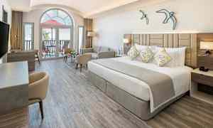 Moderni hotellihuone, jossa on leveä sänky, sohva ja työpöytä. Suuri ikkuna ja parveke taustalla. (Tekoälyn luoma teksti)