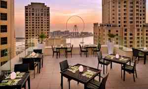 Terassi, jolla on pöytiä ja tuoleja, ympäröity korkeilla rakennuksilla, näkymä maailmanpyörään ja merelle auringonlaskun aikaan. (Tekoälyn luoma teksti)