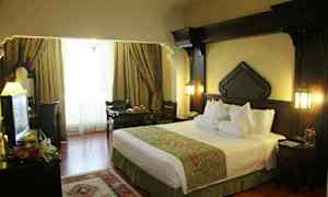 Hotellihuone, jossa on iso sänky, tummat puukalusteet ja ikkunat, joita koristavat elegantit verhot. (Tekoälyn luoma teksti)