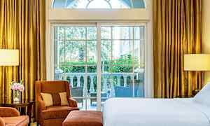 Hotellihuone, jossa suuret ikkunat avautuvat terassille ja näkymä vehreään ympäristöön. Huoneessa on sänky, nojatuoleja ja kullanväriset verhot. (Tekoälyn luoma teksti)