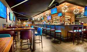 Modernin baarin sisätila, jossa on korkeita pöytiä ja tuoleja sekä useita televisioruutuja seinillä. (Tekoälyn luoma teksti)