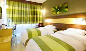 Hotellihuone, jossa on kaksi yhden hengen vuodetta, vuoteiden päätyjä koristaa piilotettu valaistus, kirjoituspöytä sekä vihreät verhot, jotka tukevat huoneen sisustusta. (Tekoälyn luoma teksti)