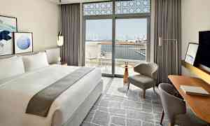 Moderni hotellihuone, jossa on suuri parivuode ja ikkunat, jotka avautuvat parvekkeelle näkymällä kaupungin siluettiin ja vedelle. (Tekoälyn luoma teksti)