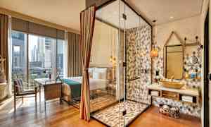 Hotellihuone, jossa on suuret ikkunat. Lasiseinäinen suihkualue näkyy pesualtaan ja peilin vieressä. (Tekoälyn luoma teksti)