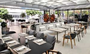 Tyylikäs ravintola, jossa on pöytiä ja istumapaikkoja, koristeltuna kasveilla ja modernilla sisustuksella. (Tekoälyn luoma teksti)